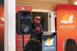 Marvis Aragon standing holding a mic behind a podium with the First Nations Logo.